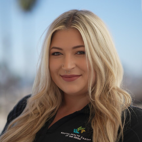 Head-and-shoulders portrait of a smiling blonde woman in a black polo with a logo, outdoors with a blue sky background.