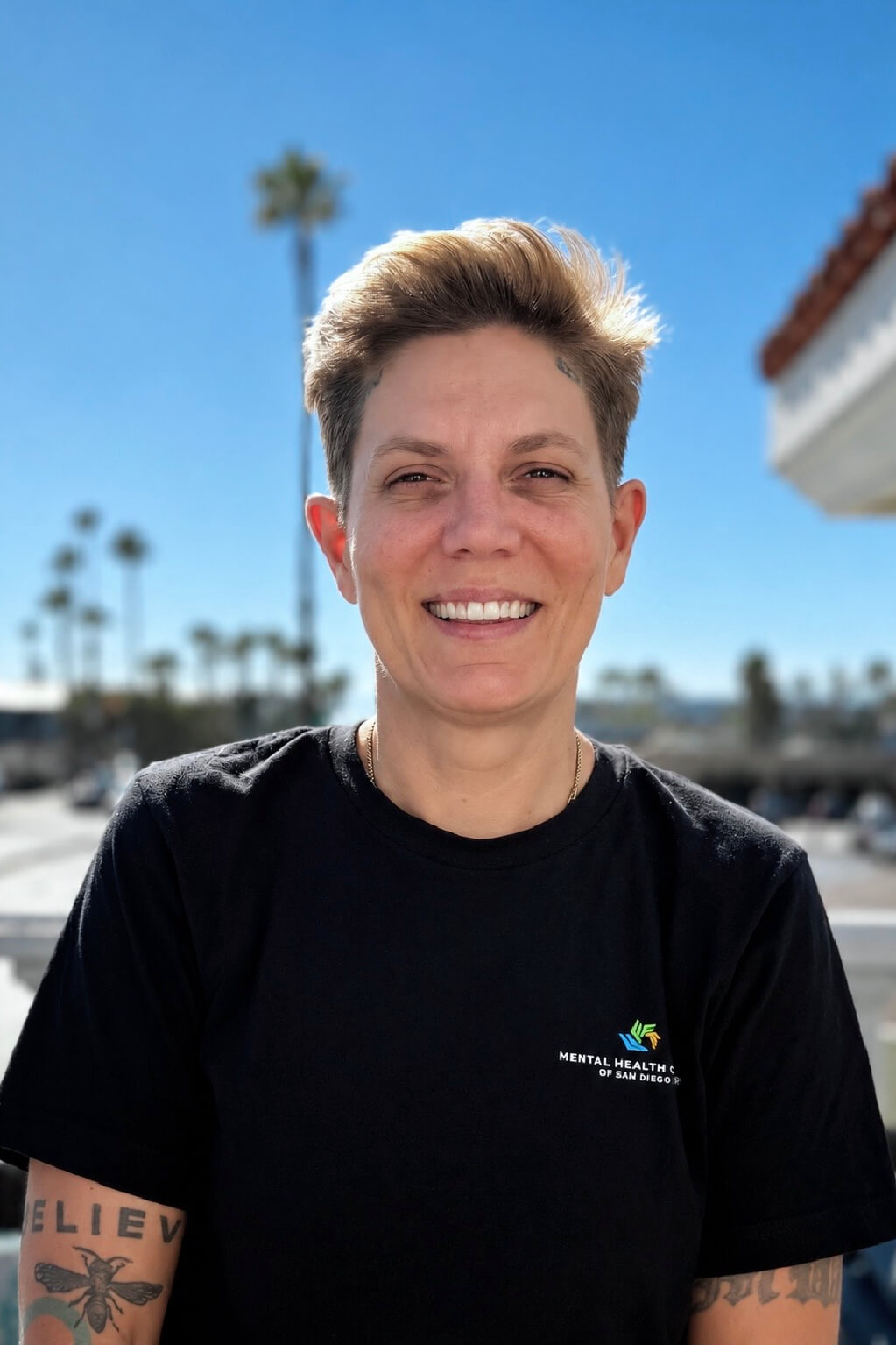 Smiling person with short hair in a black t-shirt outdoors, blue sky and palm trees in the background.