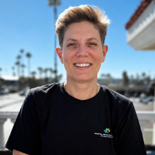 Smiling person with short hair wearing a black shirt, outdoors on a sunny day with palm trees in the background.