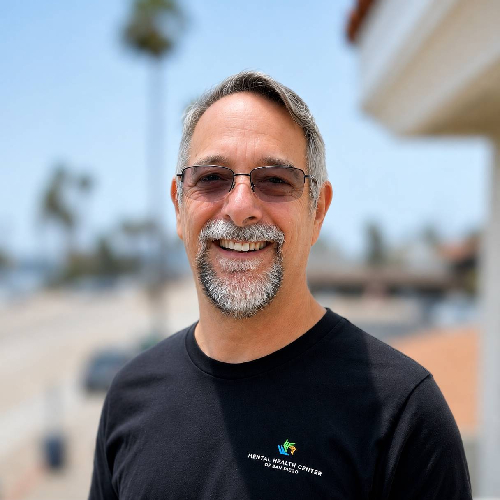 Smiling man with gray hair and goatee wearing sunglasses, standing outdoors on a sunny day with palm trees in the background.