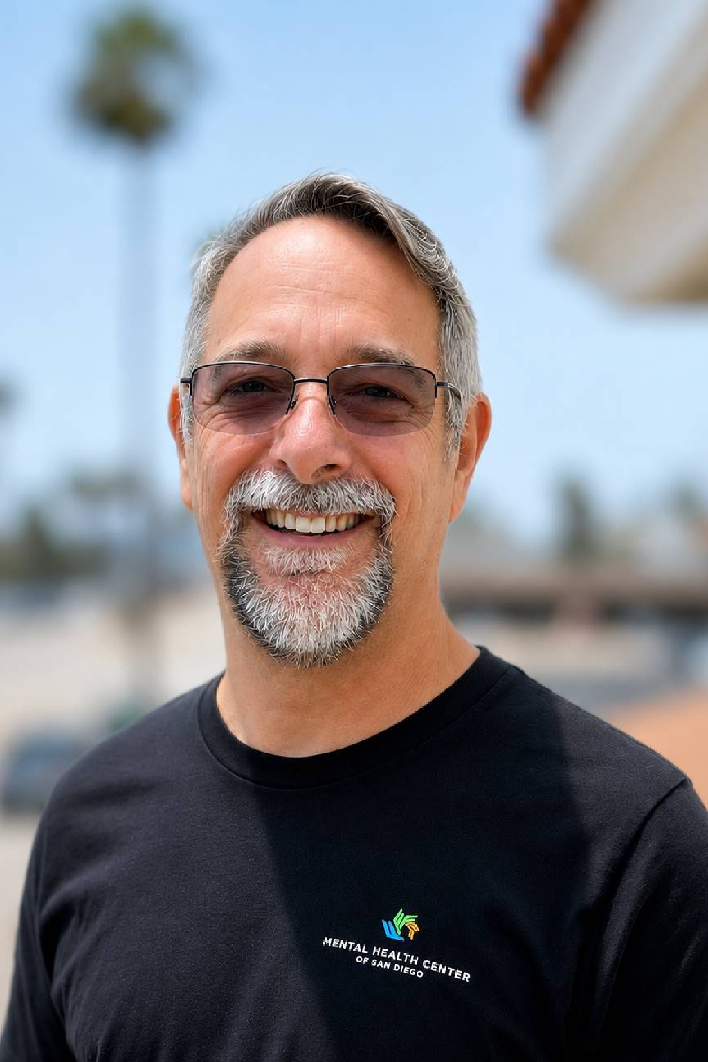 Smiling middle-aged man with gray hair and beard, wearing sunglasses and a black T-shirt with a Mental Health Center of San Diego logo, outdoors.