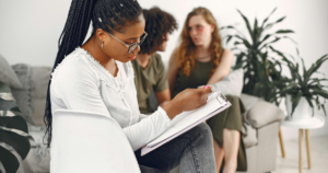 Woman with glasses writes on a clipboard during a group therapy or counseling session, with two peers conversing in the background.