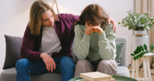 A man sits beside a distressed woman on a sofa, offering support with his hand on her shoulder in a cozy living room.