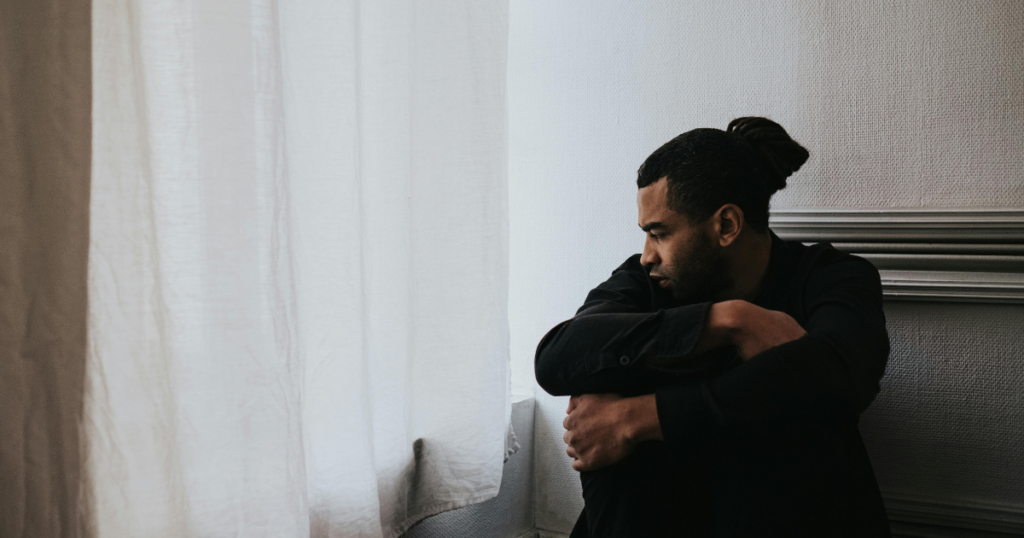 Man sitting on the floor by a window, hugging his knees and looking down, appears thoughtful or sad.