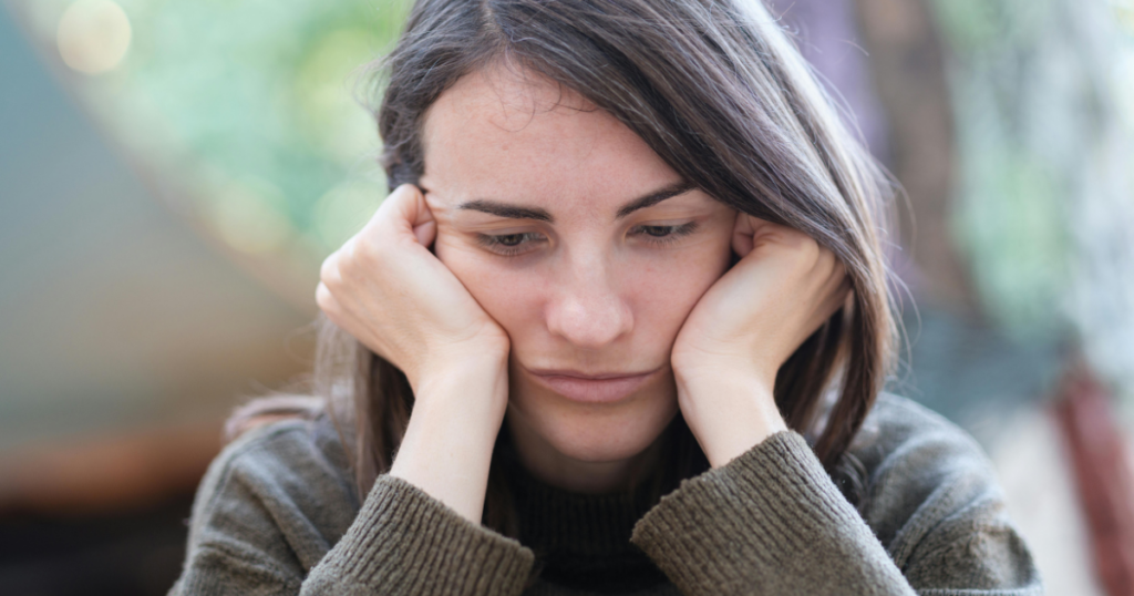 Young woman with brown hair resting her face on her hands, looking sad or pensive outdoors in a soft-focus setting.