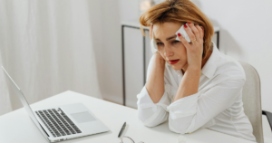 Woman at a white desk with a laptop, looking stressed and wiping her face with tissues while seated in a white shirt.