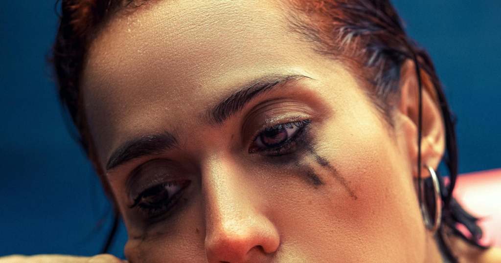 image - Mental Health Center Of San Diego Close-up of a person with smudged black eye makeup under the eye, looking downward against a blue background, wearing a hoop earring (portrait).