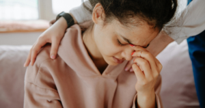 Young woman in a pink hoodie sits with eyes closed as a friend places a comforting arm around her shoulder.