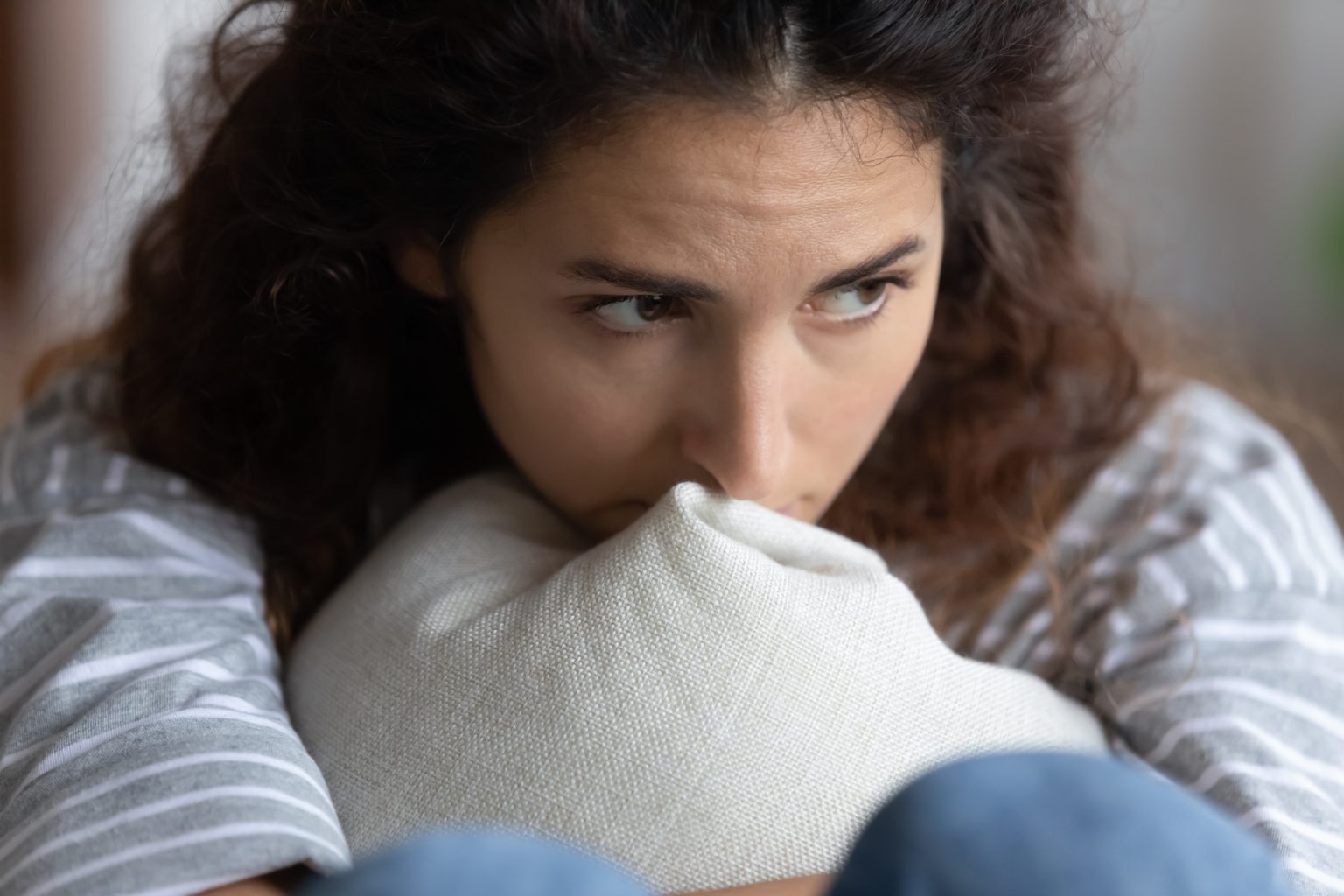 Woman sitting with a pensive, worried expression while hugging a light pillow for comfort indoors.