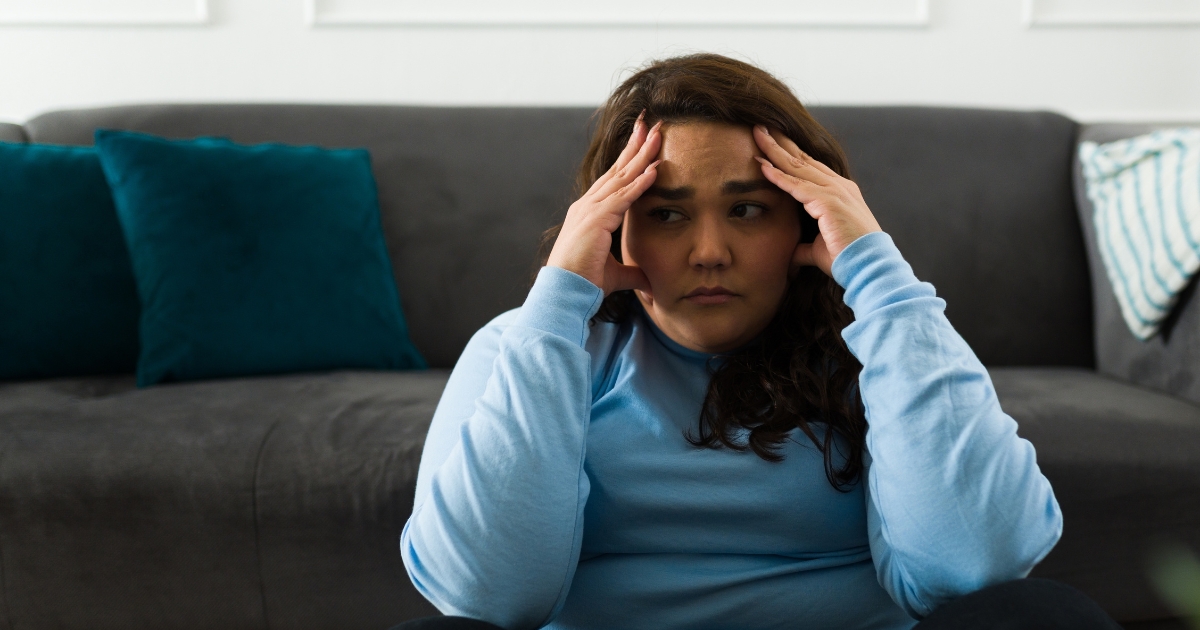 Woman sitting on the floor in a blue long-sleeve shirt, hands on her temples with a worried expression on a gray sofa backdrop