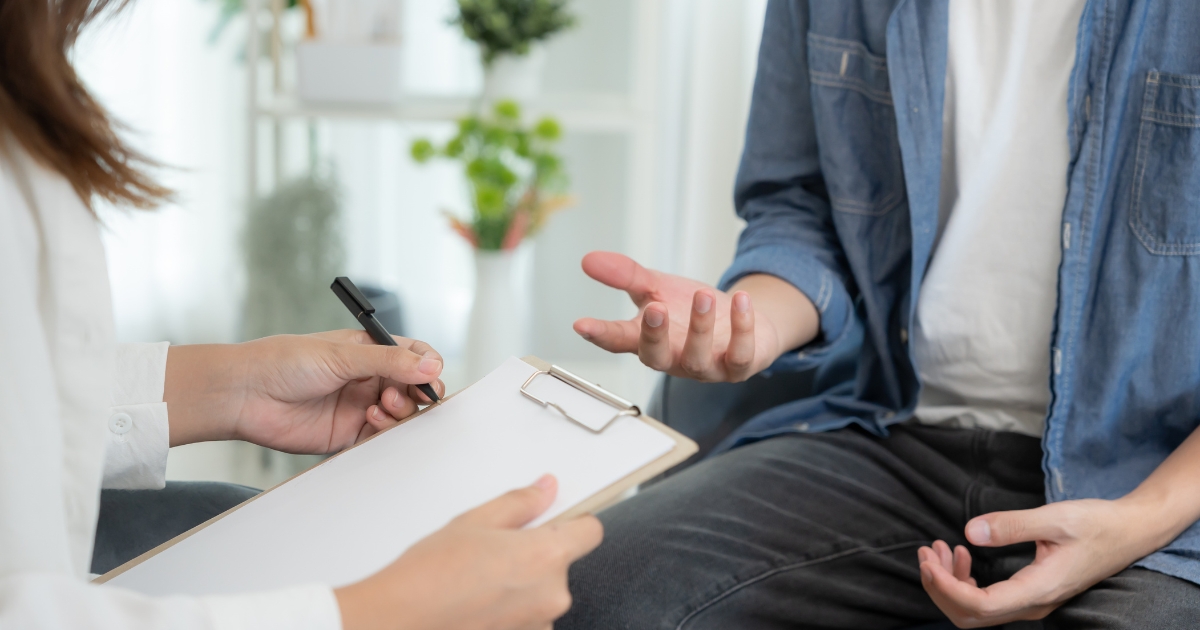 Clinician writes on a clipboard while a patient gestures with hands during a consultation.
