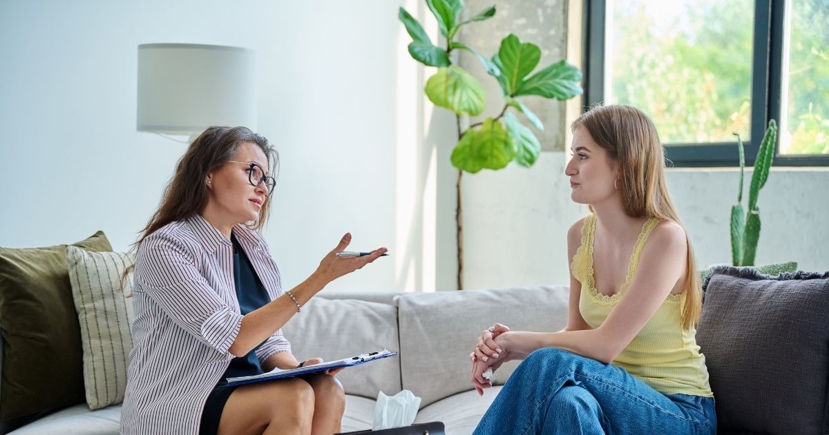 A woman therapist sits on a sofa with a clipboard and pen, talking to a young woman seated opposite her in a bright living room setting.