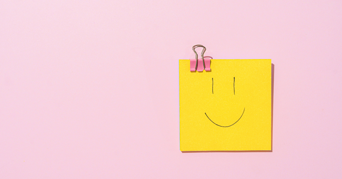 Yellow sticky note with a smiley face clipped to a pink binder clip on a pink background.