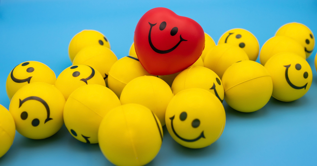 Red smiling heart stands out among a pile of yellow smiley balls on a blue background with a cheerful vibe.