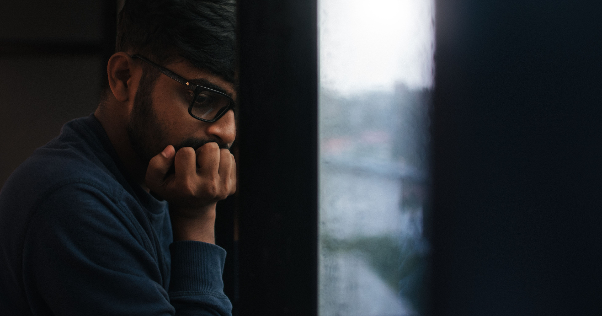 Young man with glasses sits by a window, resting his chin on his hands and looking down in thought.