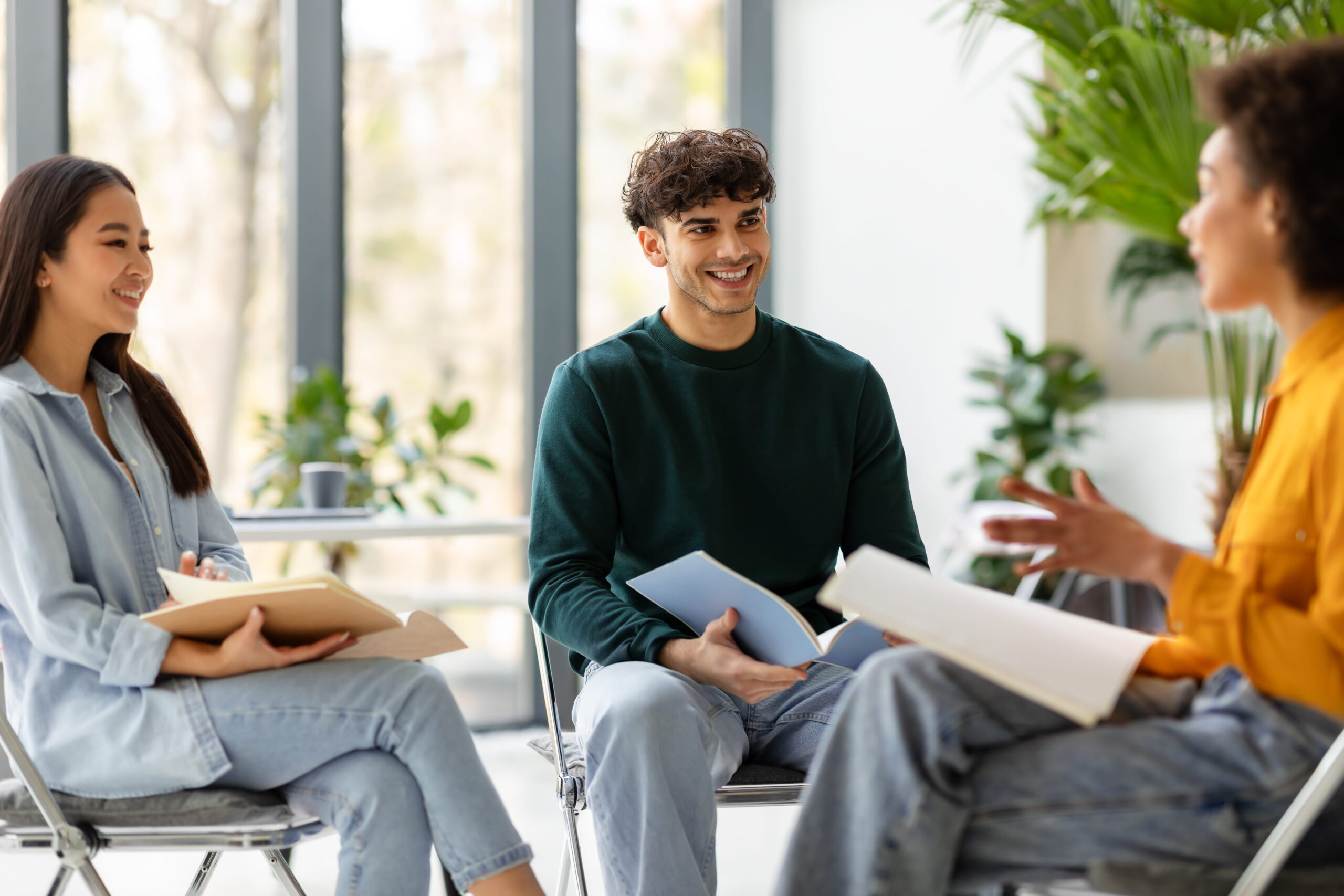 Three people sit in a bright room, smiling and talking while holding notebooks and papers during a group discussion.