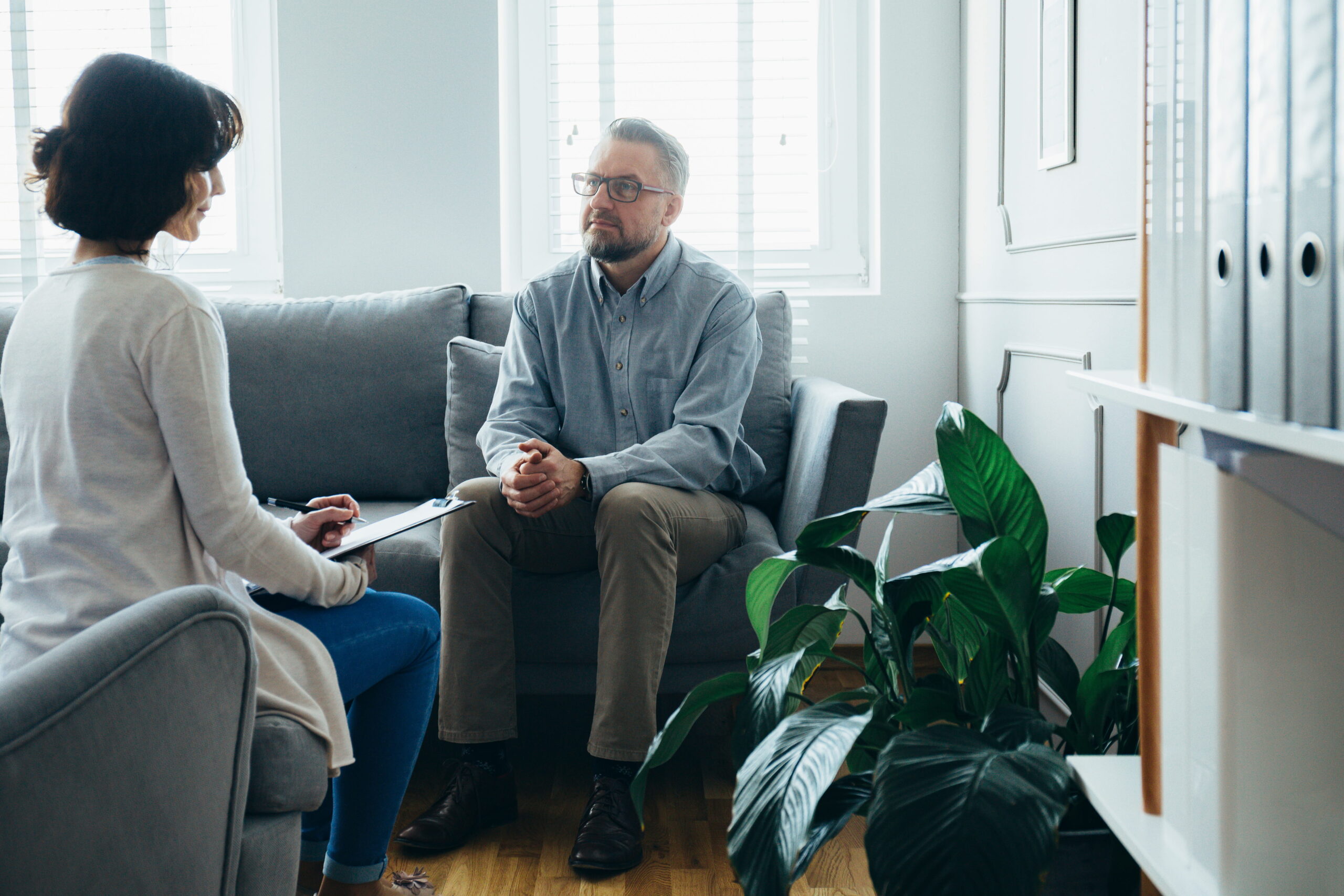Therapist takes notes on a clipboard while a man with glasses sits on a gray sofa in a bright office.