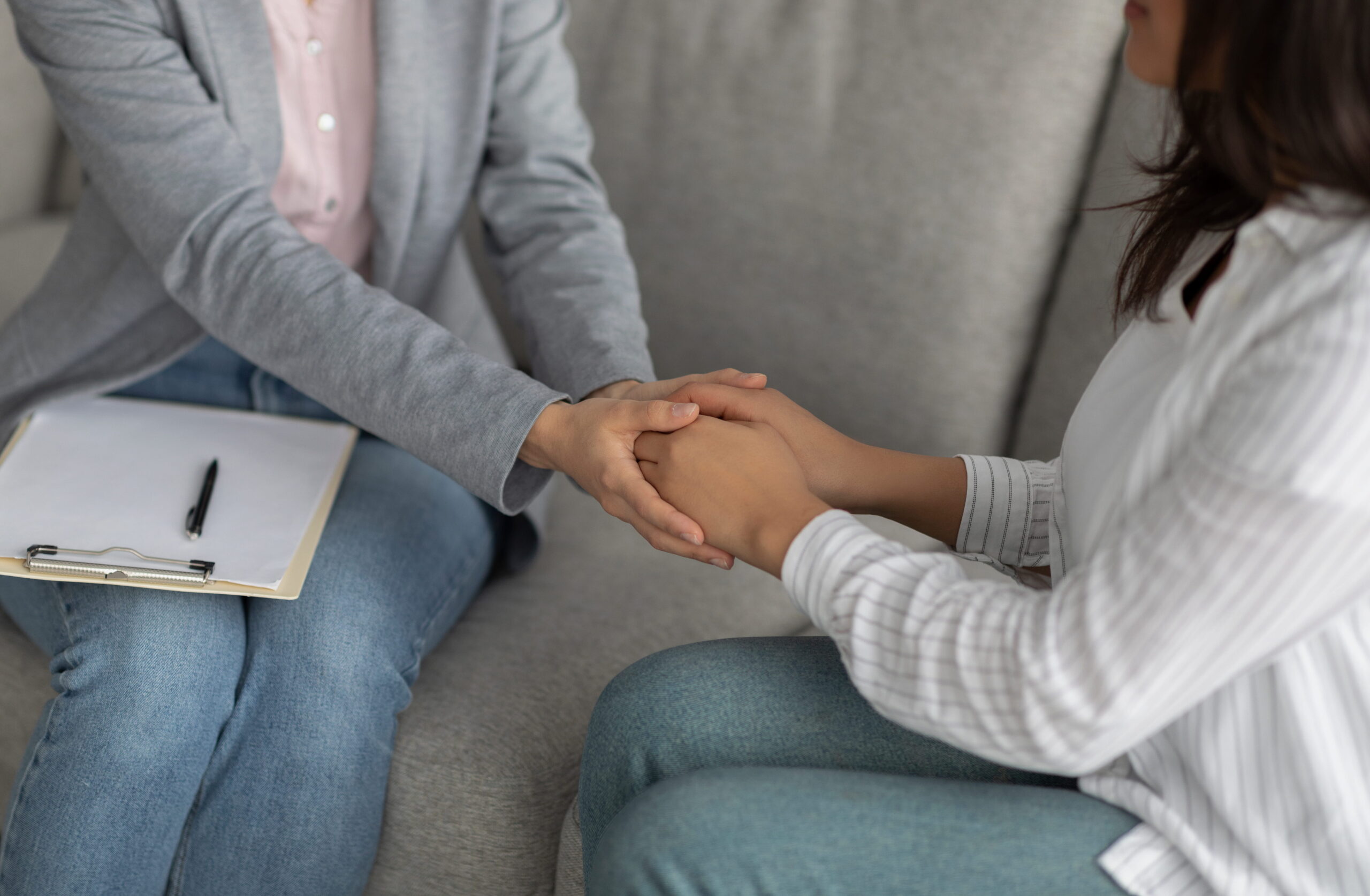 Two people sit together, one holding the other's hands in a comforting gesture, with a clipboard nearby.