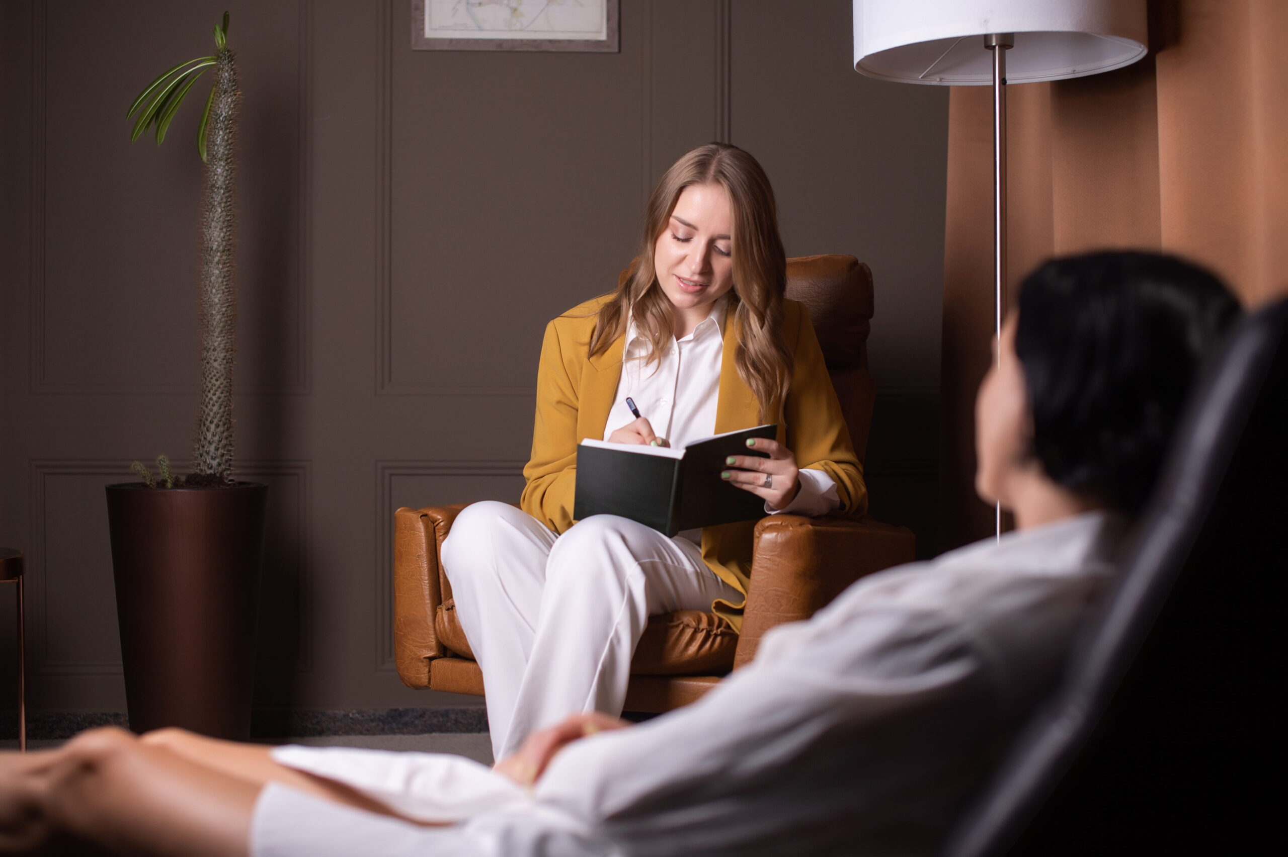 Woman in a mustard blazer writes in a notebook while seated in a brown leather chair in an office setting, with a blurred person in the foreground.
