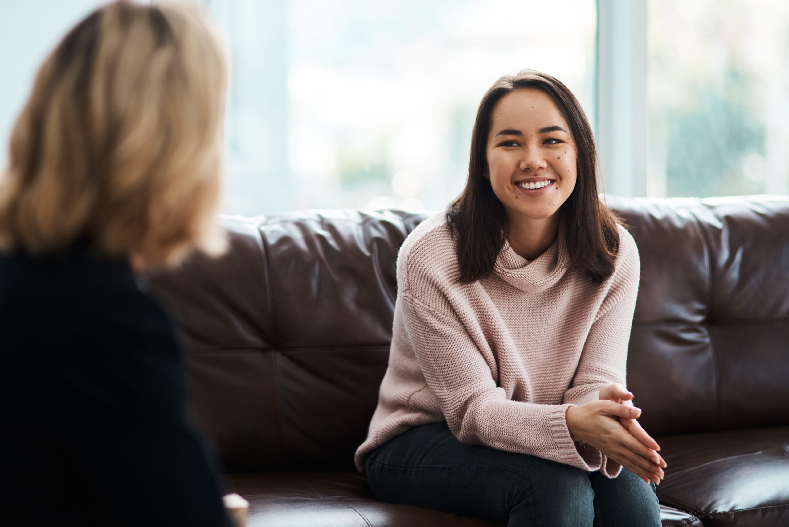 Smiling woman in a pink sweater sits on a brown leather couch, talking with a blurred person in the foreground.